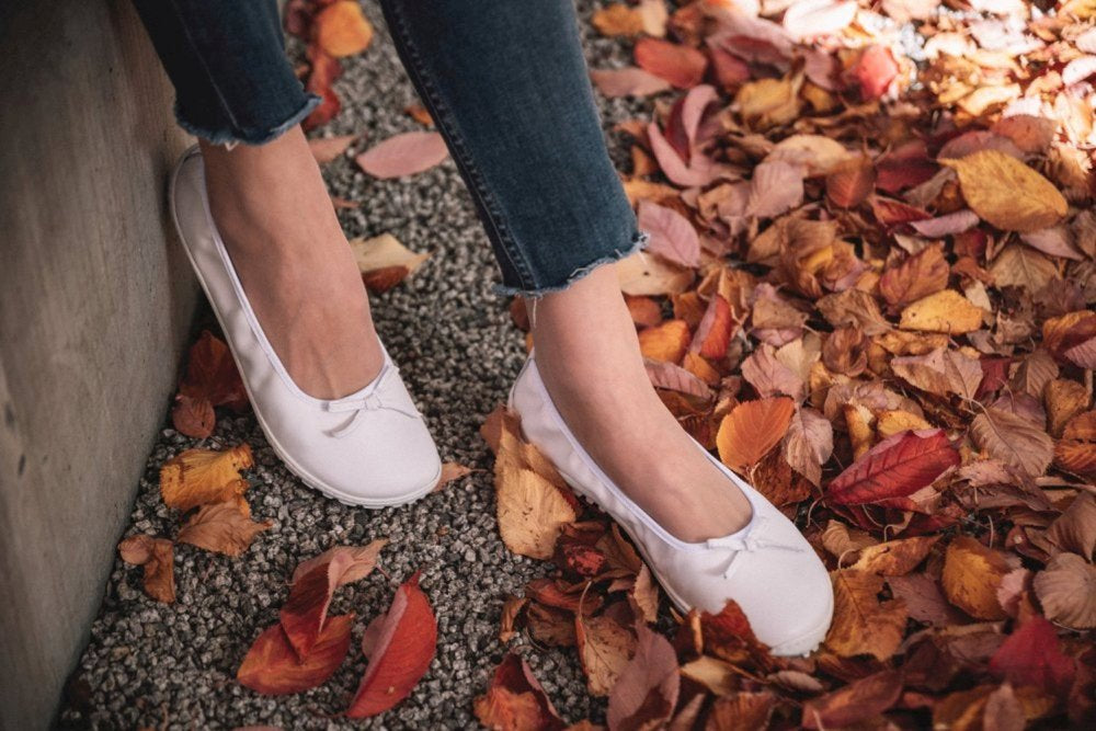 Eine Person steht auf einem Kiesweg, umgeben von gefallenen Herbstblättern in Rot-, Orange- und Brauntönen und trägt die weißen Ballerinas SUQAR mit Sneakersohle von ZAQQ und dazu dunkle Jeans.