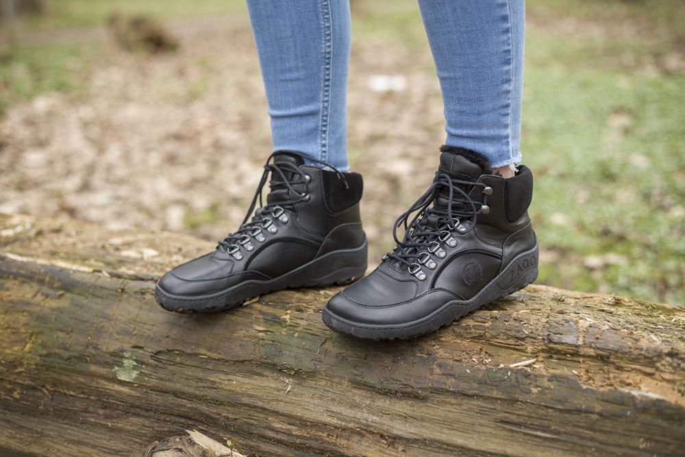 In den schwarzen wasserdichten Winterstiefeln ZAQQ TREQ und Blue Jeans steht eine Person selbstbewusst auf einem großen Baumstamm inmitten einer Waldlandschaft. Der Boden ist mit Blättern bedeckt und bietet die perfekte Kulisse für ein Outdoor-Abenteuer.