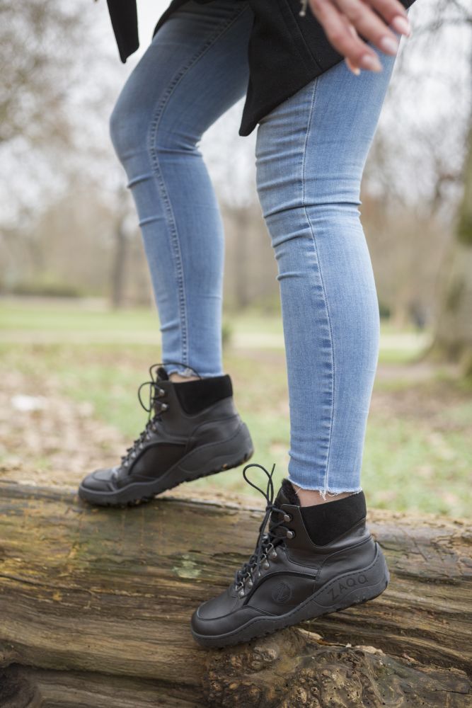 Eine Person in den wasserdichten schwarzen TREQ Winterstiefeln von ZAQQ und hellblauen Jeans balanciert auf einem großen Baumstamm in einer Außenkulisse mit verschwommenen Bäumen und Gras im Hintergrund.