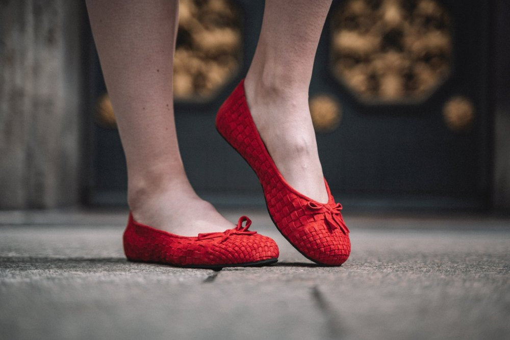 Eine Person in den TWIST Red Barfuß-Ballerinas mit Schleifchen von ZAQQ steht auf einem Steinboden. Der dunkle Untergrund ist mit einem dekorativen Muster versehen, welches die kräftige Farbe der Schuhe hervorhebt.