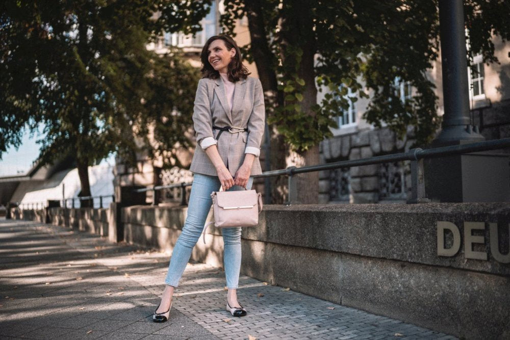 Eine Frau in einem grauen Blazer und Jeans steht lächelnd auf einem von Bäumen gesäumten Gehweg. Sie hält ihre TWIST Varnish Nude Handtasche von ZAQQ aus weichem Nappaleder in der Hand und scheint den sonnigen Tag zu genießen. Im Hintergrund sind ein Gebäude und eine Steinmauer zu sehen, die ihren heiteren Moment noch verstärken.