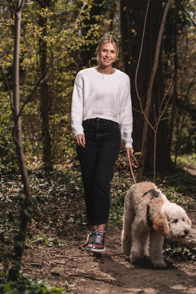 Eine Person spaziert mit einem flauschigen Hund an der Leine durch einen Waldweg. Sie trägt einen weißen Pullover, schwarze Hosen und leuchtend rote VAQATION-Sneaker von ZAQQ. Das Sonnenlicht fällt durch die Bäume und beleuchtet den Weg, während sie den Komfort ihrer wasserdichten Schuhe genießt.